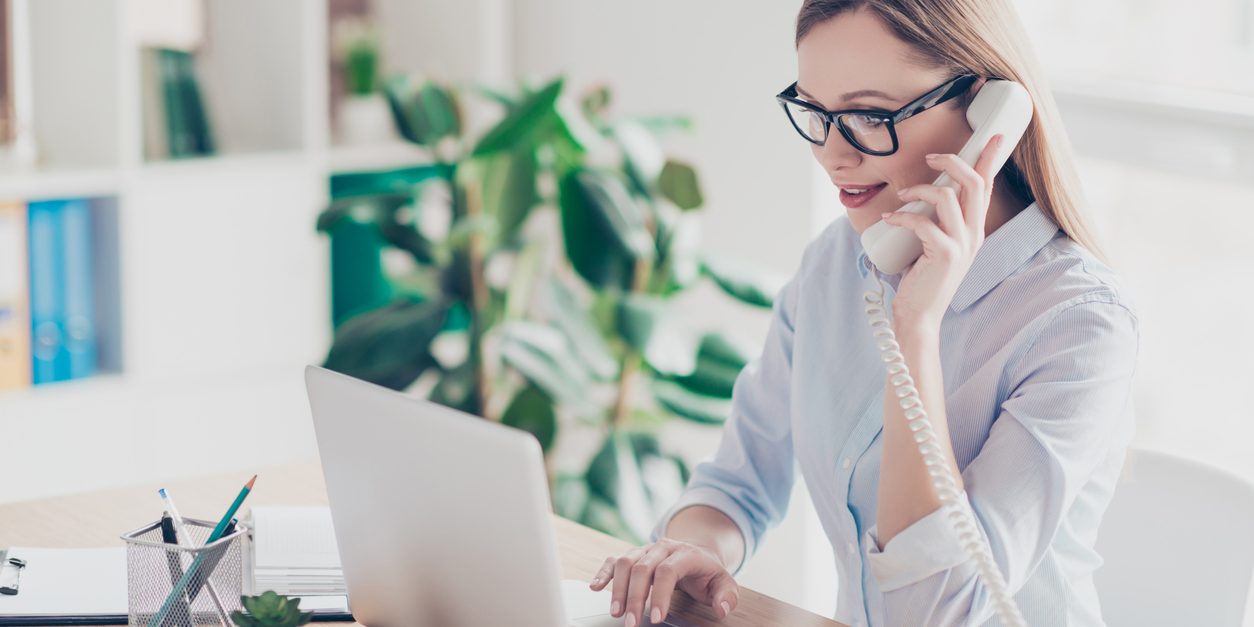 Portrait of busy, cheerful, attractive, pretty operator with hairstyle holding handset near ear, speaking on phone, checking information on pc, sitting in work place, station at desktop on chair va